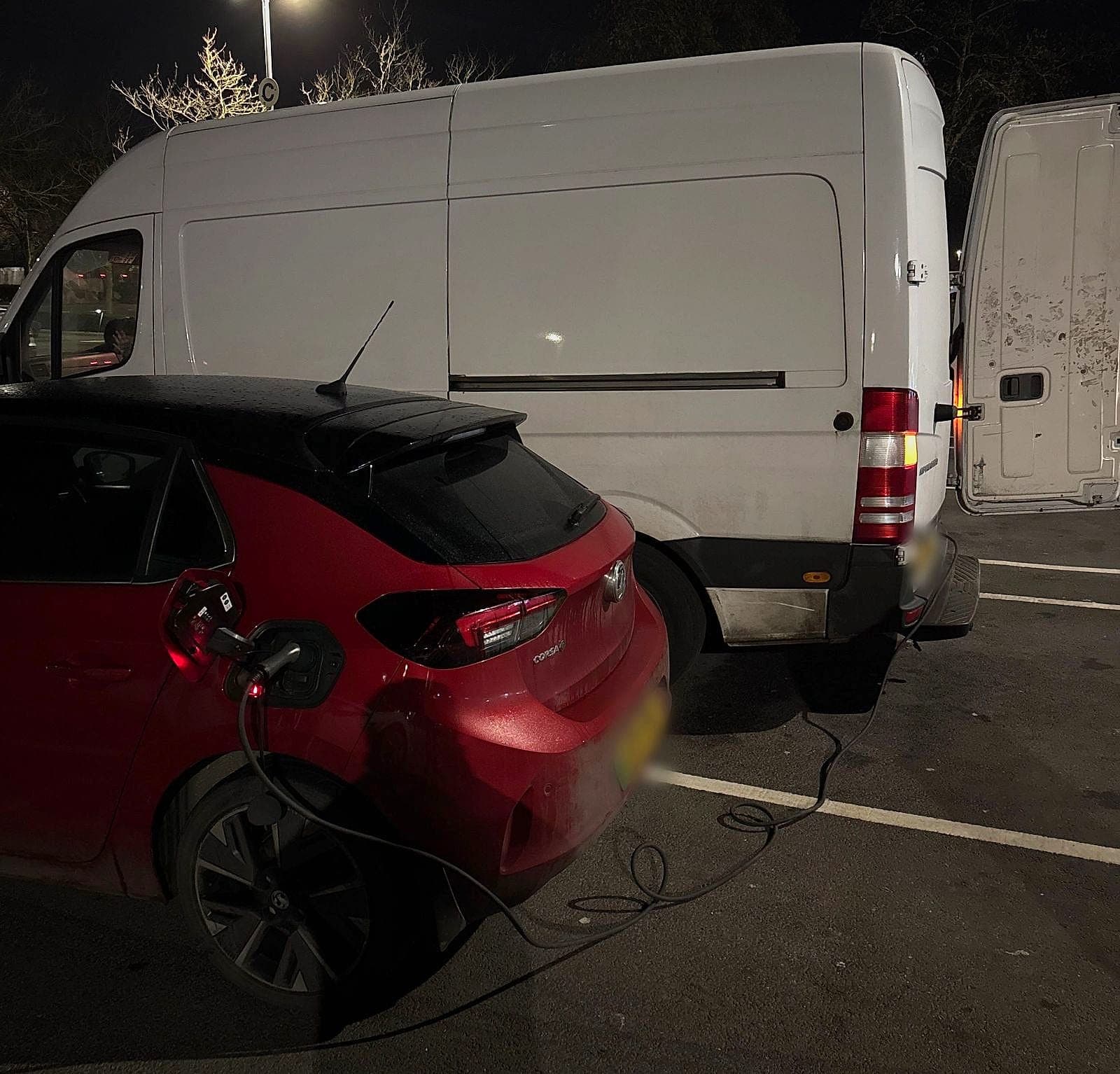 A red Vauxhall Corsa Electric charging in a car park at night beside a white van