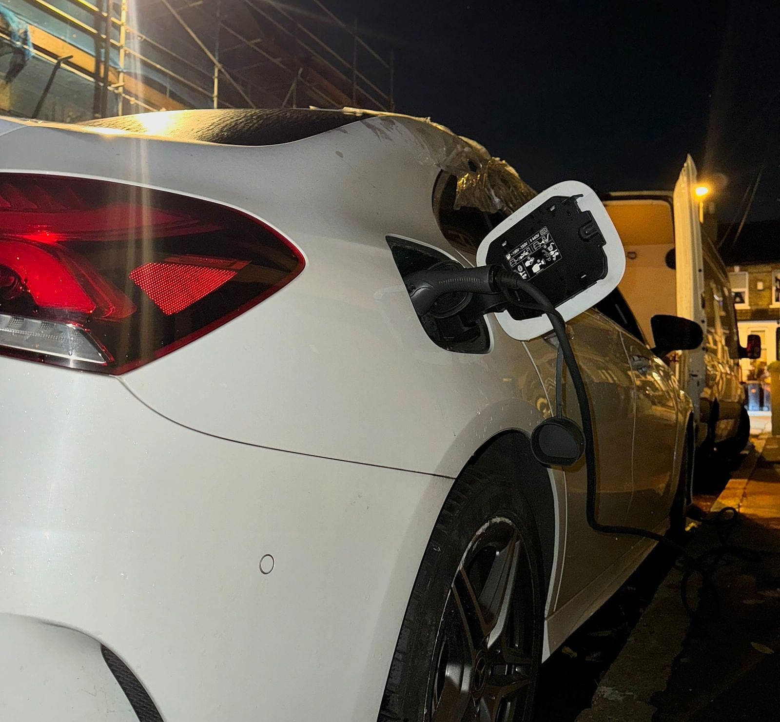Close up of a white car's open charge flap with the connector plugged in at night